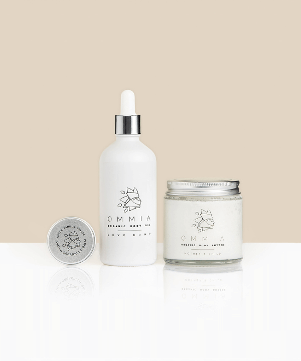 The image displays the OMMIA Love Mom Bundle, featuring three premium skincare products: the OMMIA Organic Lip Balm in a sleek silver tin, the OMMIA Organic Body Oil in a clean white dropper bottle labeled "Love Bump," and the OMMIA Organic Body Butter in a glass jar labeled "Mother & Child." All products are elegantly arranged against a soft beige background, showcasing their minimalist, natural aesthetic. Perfect for nurturing and pampering mothers with organic care.